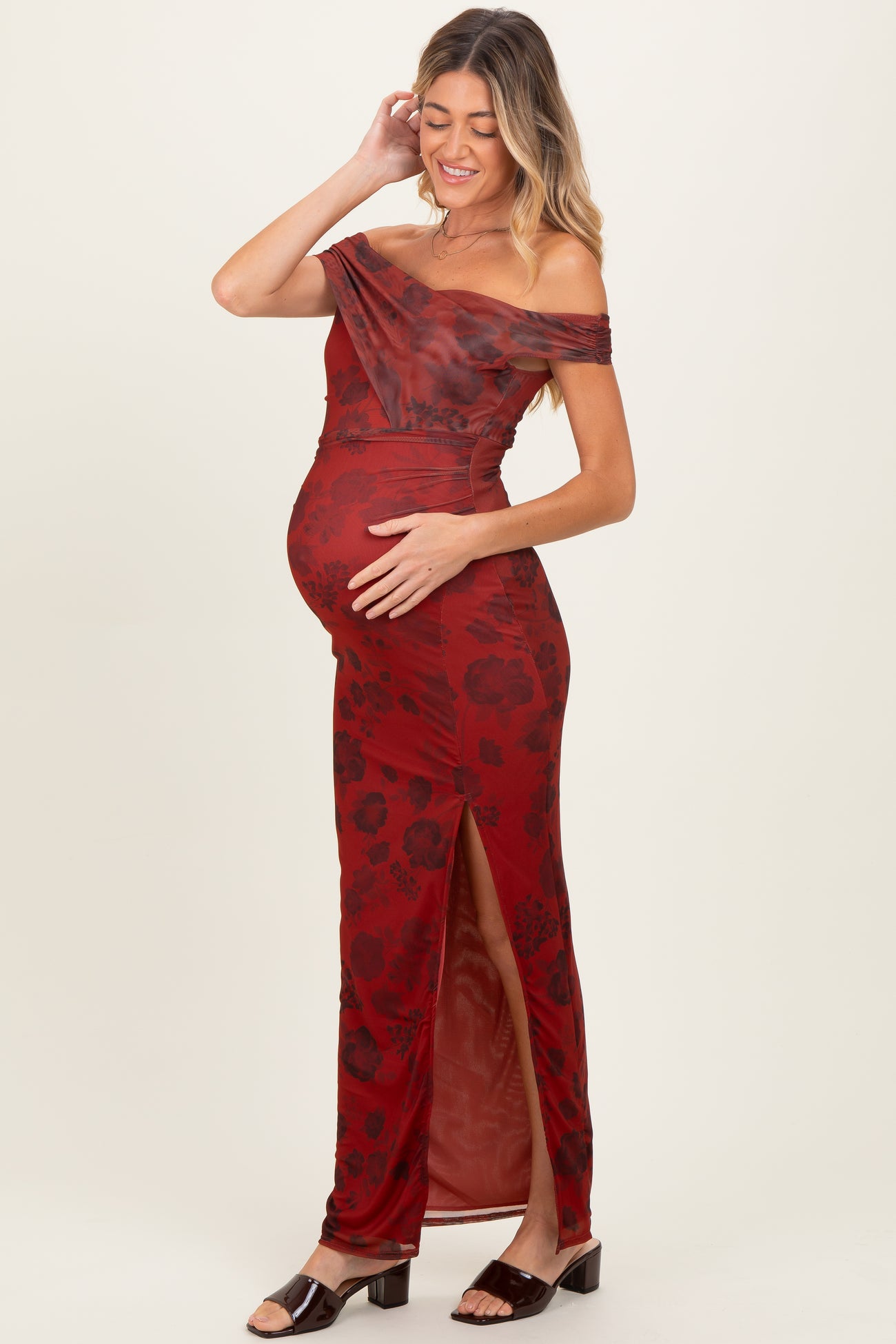 woman wearing a red floral off-the-shoulder maternity gown with a side slit and holding her baby bump