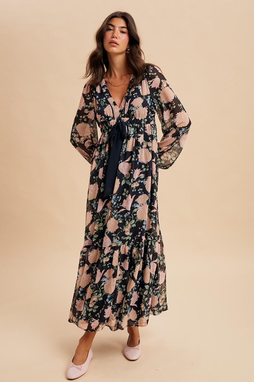 woman wearing a long floral maxi dress with a tie detail and bell sleeves, paired with pink flats