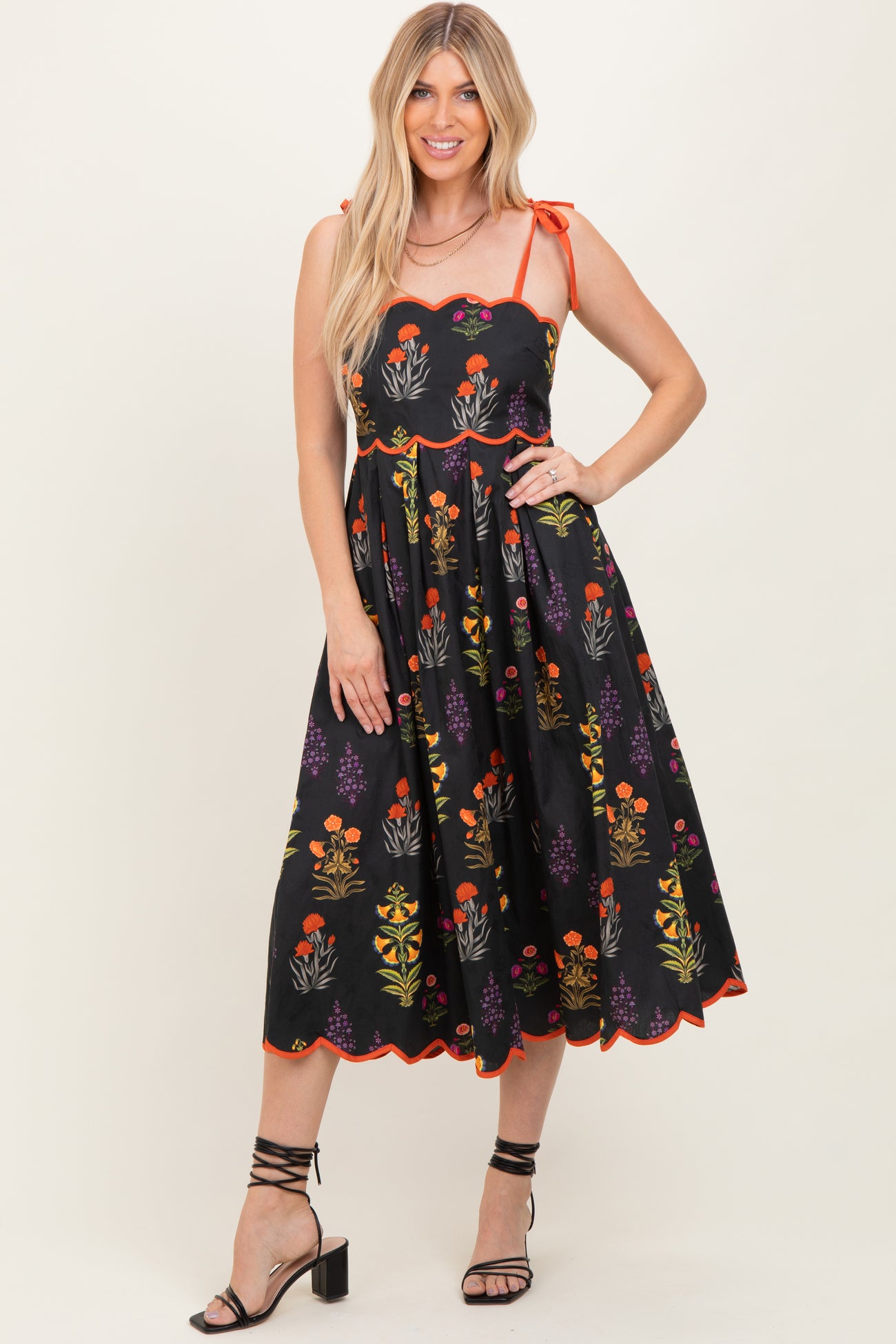 woman wearing a black floral midi dress with tie straps and scalloped edges, standing with one hand on her hip