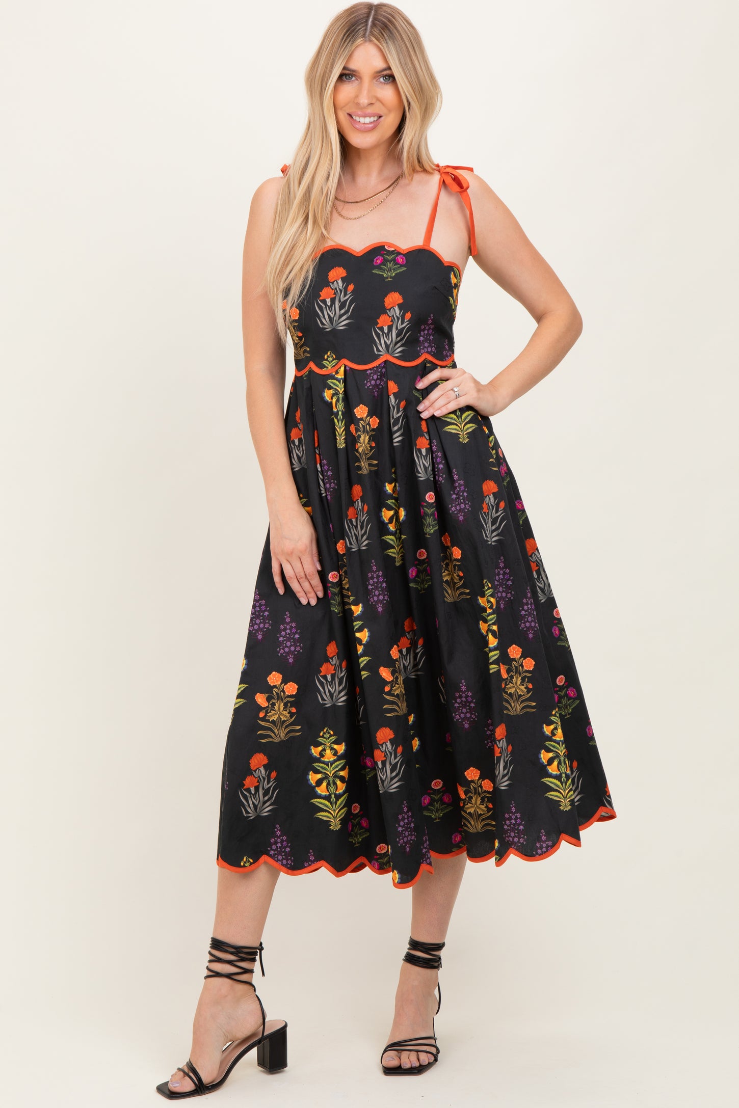 woman wearing a black floral midi dress with tie straps and scalloped edges, standing with one hand on her hip