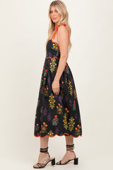woman wearing a black embroidered sundress with colorful floral designs and orange accents, standing sideways