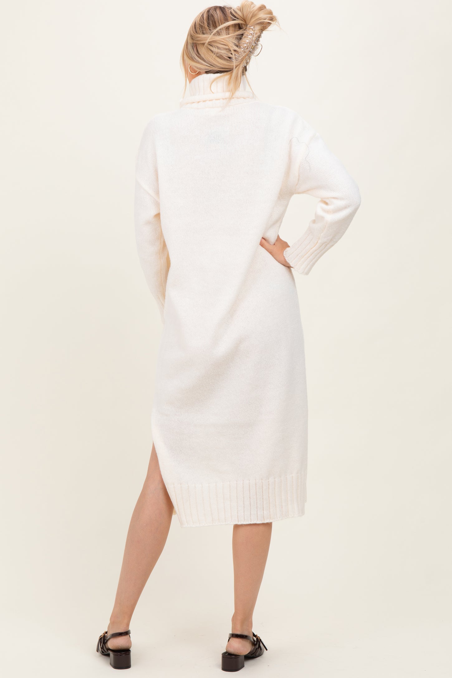 woman wearing a cream-colored long sleeve sweater dress with a high collar and side slit, viewed from the back