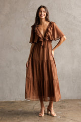 woman wearing a brown tiered maxi dress with ruffled sleeves and a tie waist, standing against a textured wall