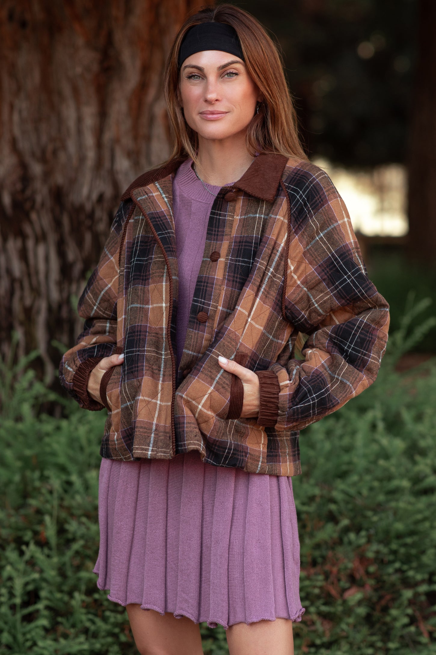 woman wearing a brown plaid cropped jacket over a purple pleated dress with a headband in her hair