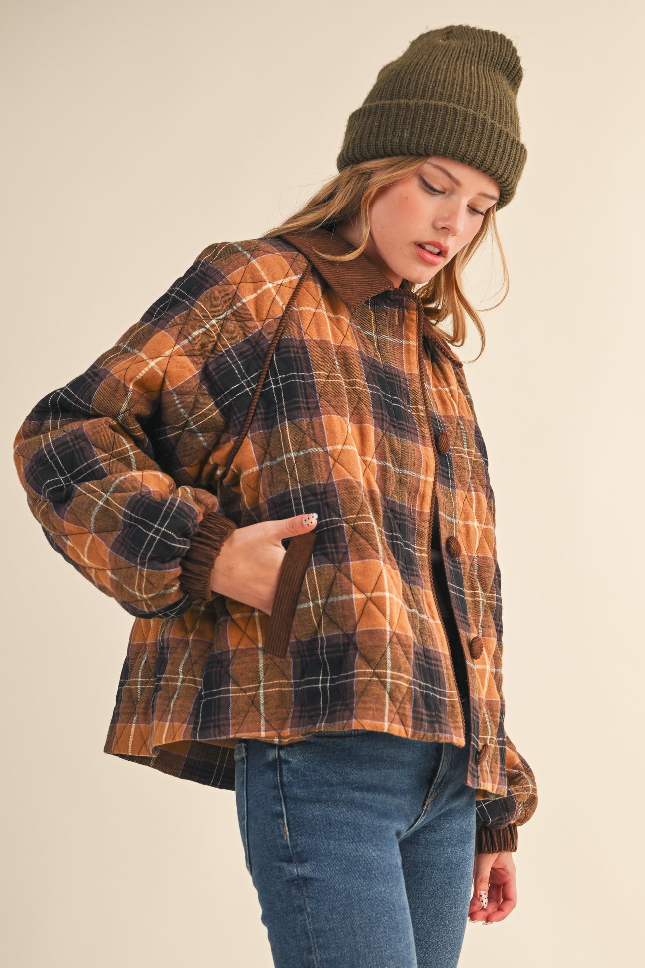 woman wearing a brown and navy plaid quilted jacket with a hood and a knit beanie, styled with jeans