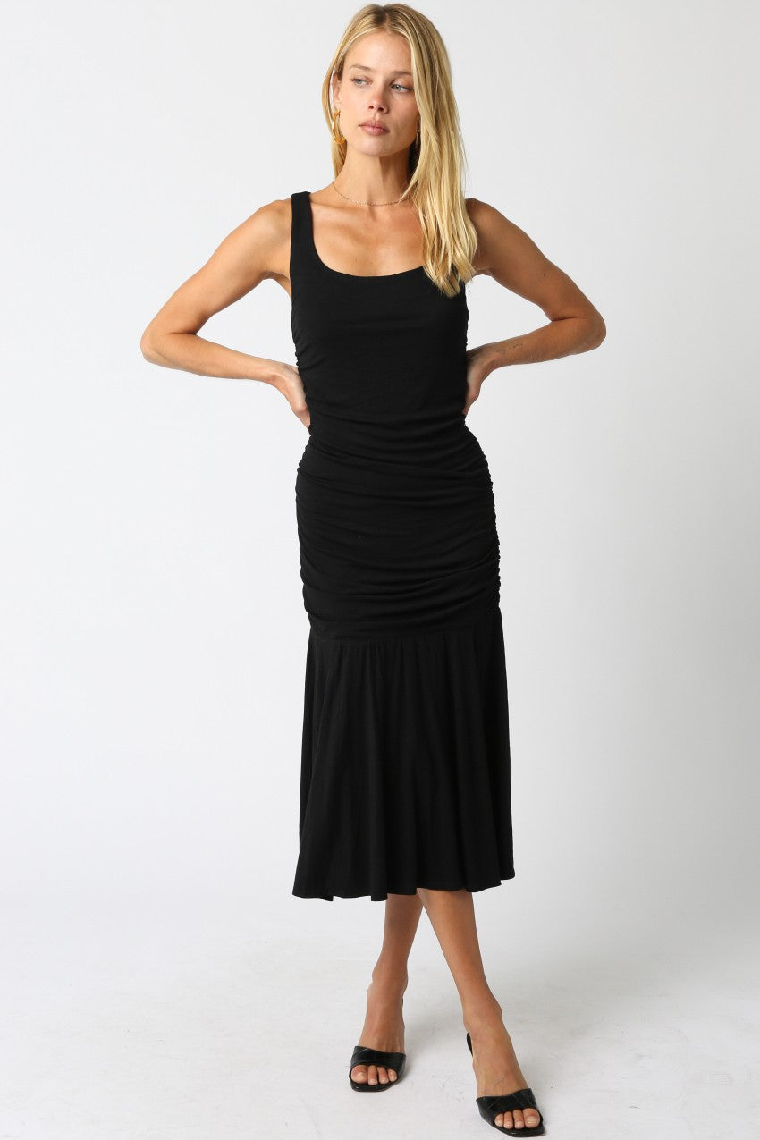 woman wearing a black fitted midi dress with ruching detail and a flared hem, posing with hands on hips