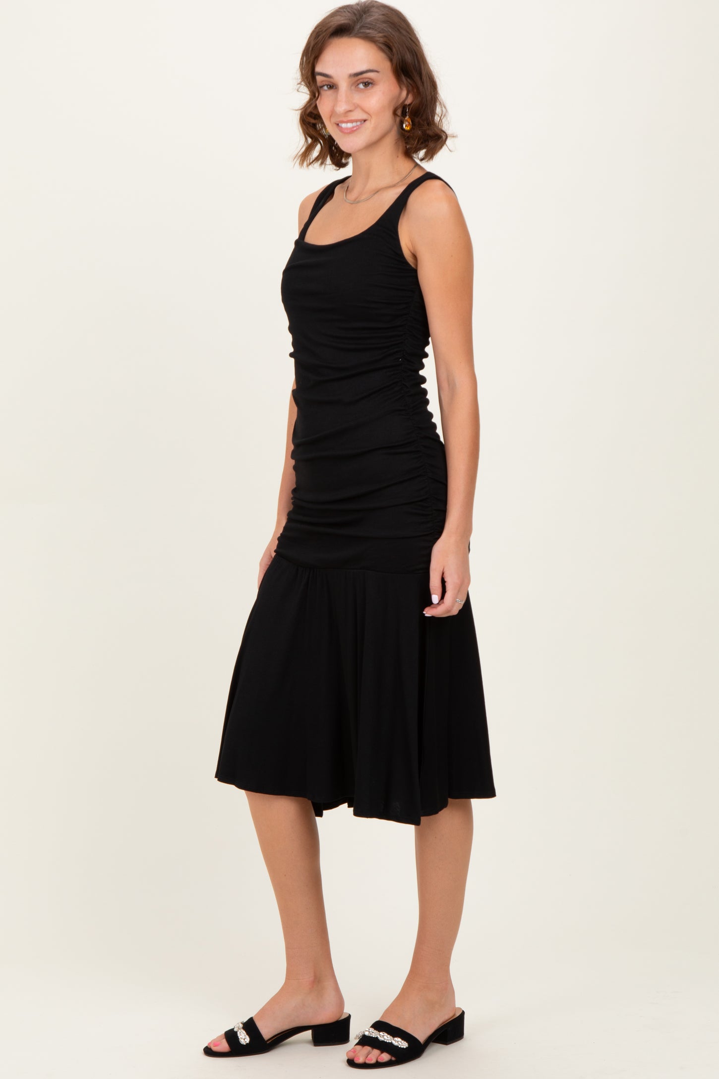 woman wearing a black sleeveless ruched dress with a flared skirt and black slide sandals