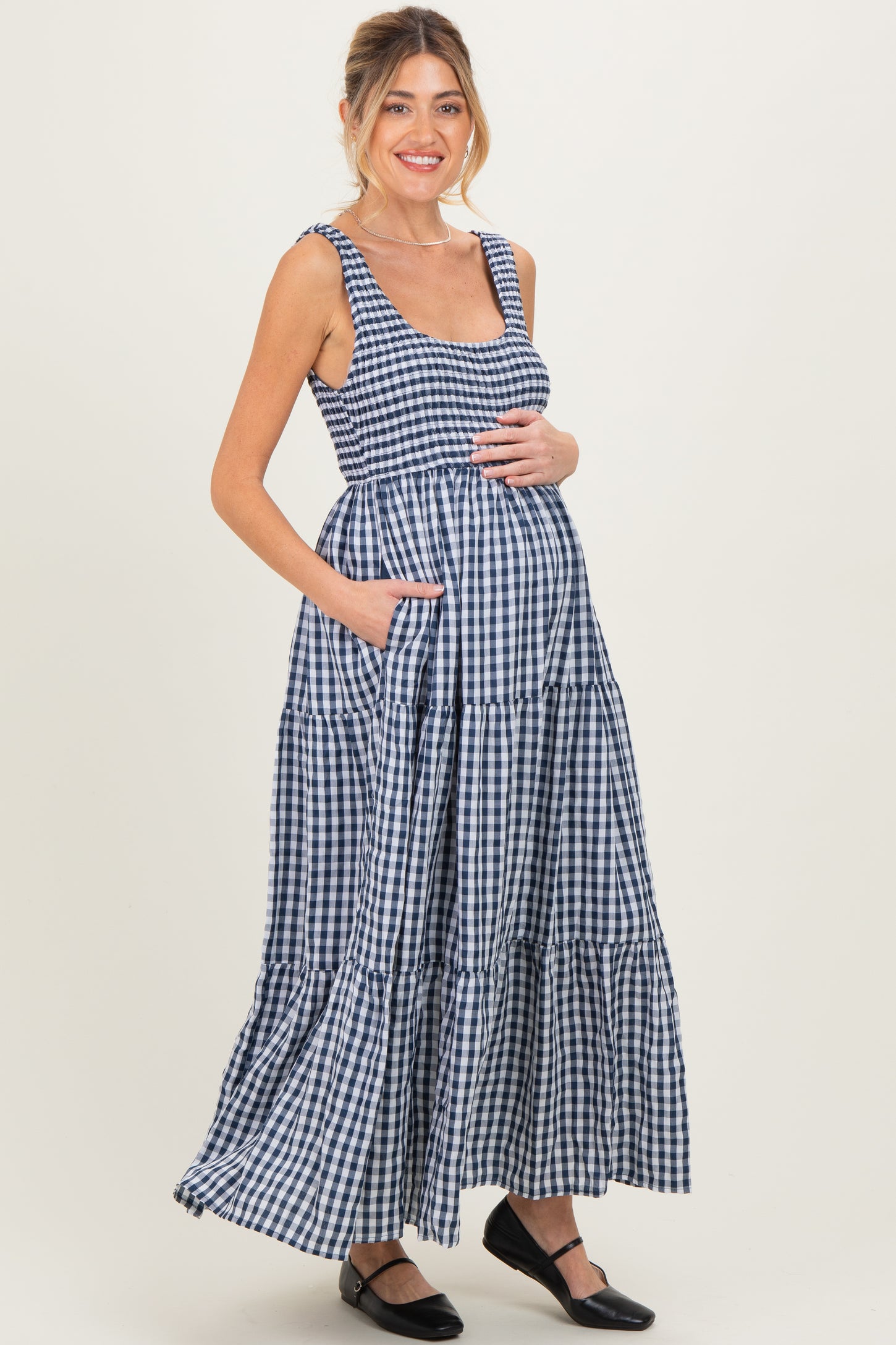 woman wearing a navy and white gingham maternity maxi dress with a tiered skirt and pockets, highlighting her baby bump