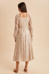 woman wearing a beige textured long sleeve dress with floral embroidery and a fitted waist, viewed from the back