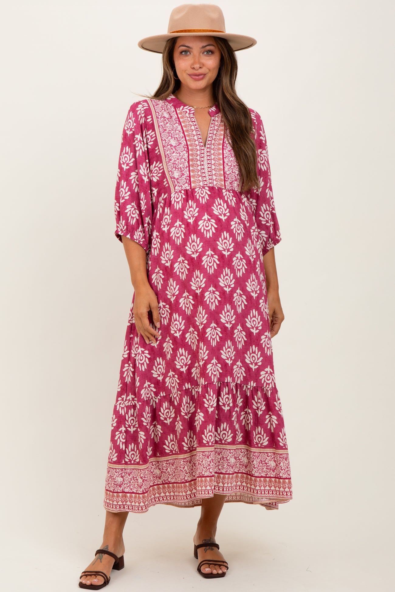 woman wearing a pink and white floral print maternity maxi dress with bell sleeves and a v-neckline, smiling and posing with a hat