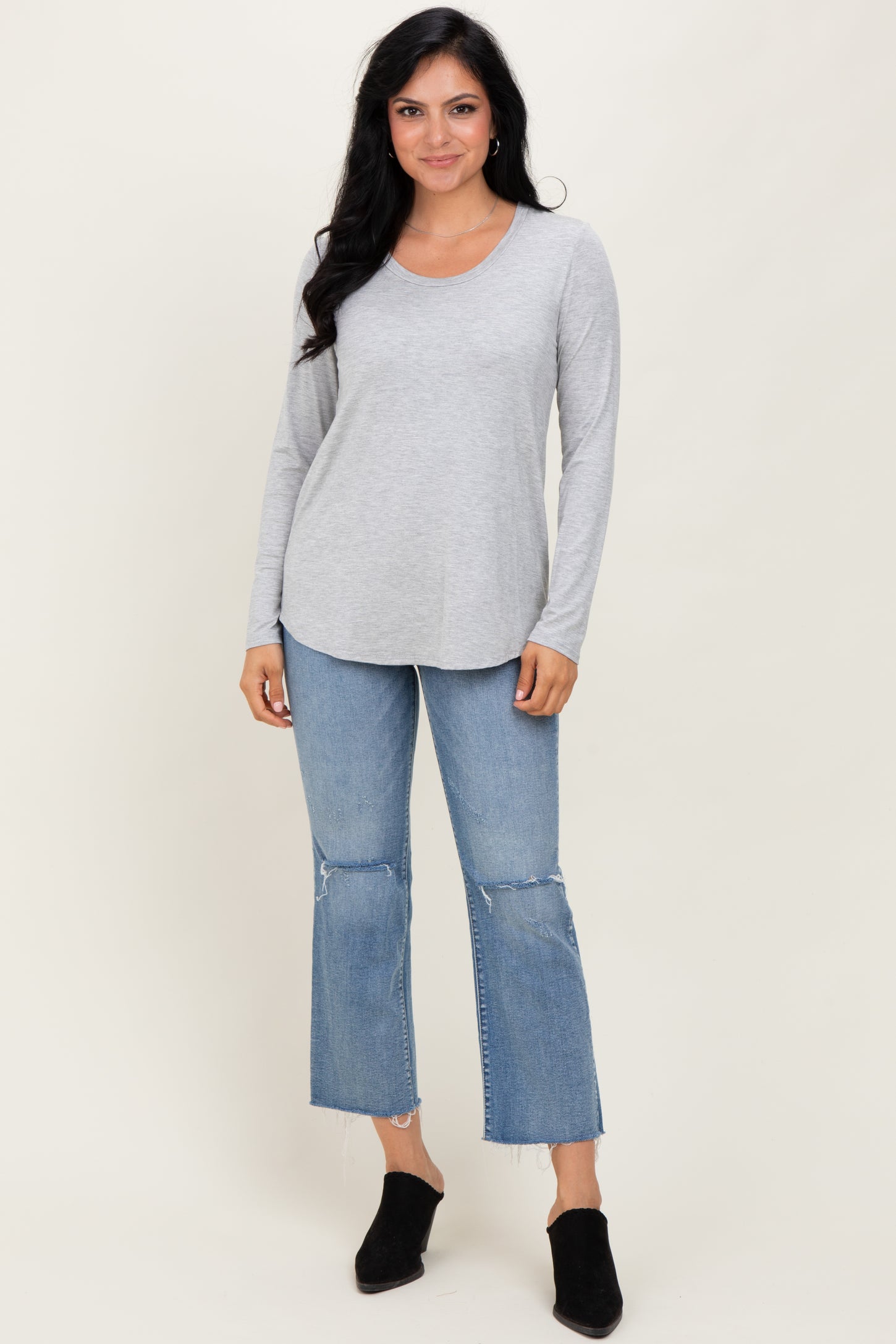 woman wearing a light gray long sleeve top with a curved hem paired with distressed blue jeans and black ankle boots
