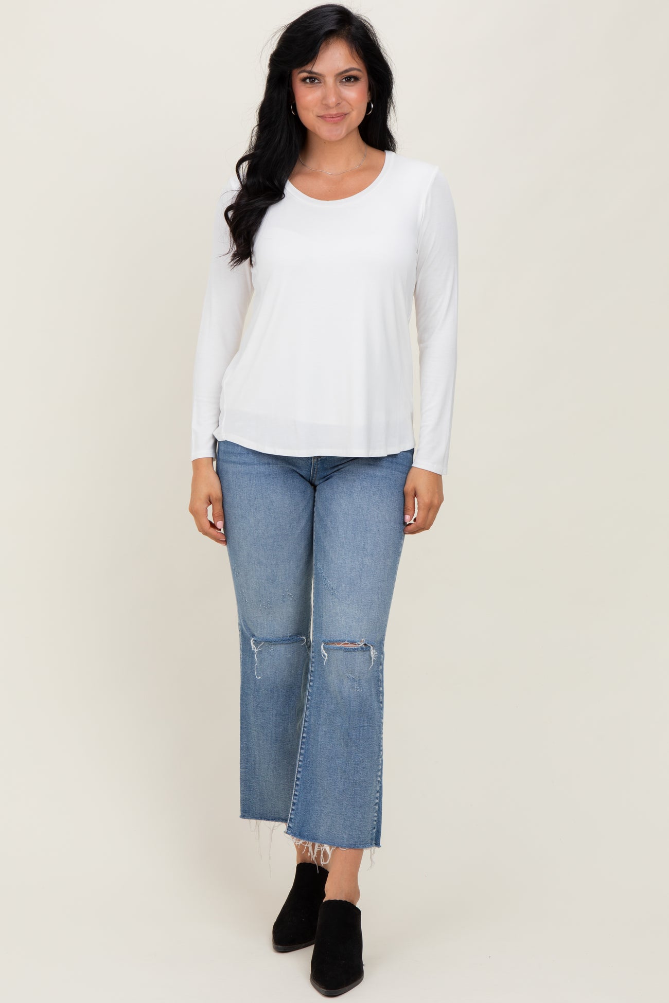woman wearing a white long sleeve top with scoop neckline and light blue ripped jeans, standing against a neutral background