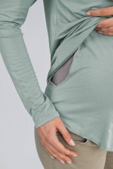 woman wearing a soft green long sleeve maternity top with side pockets, gently holding her baby bump