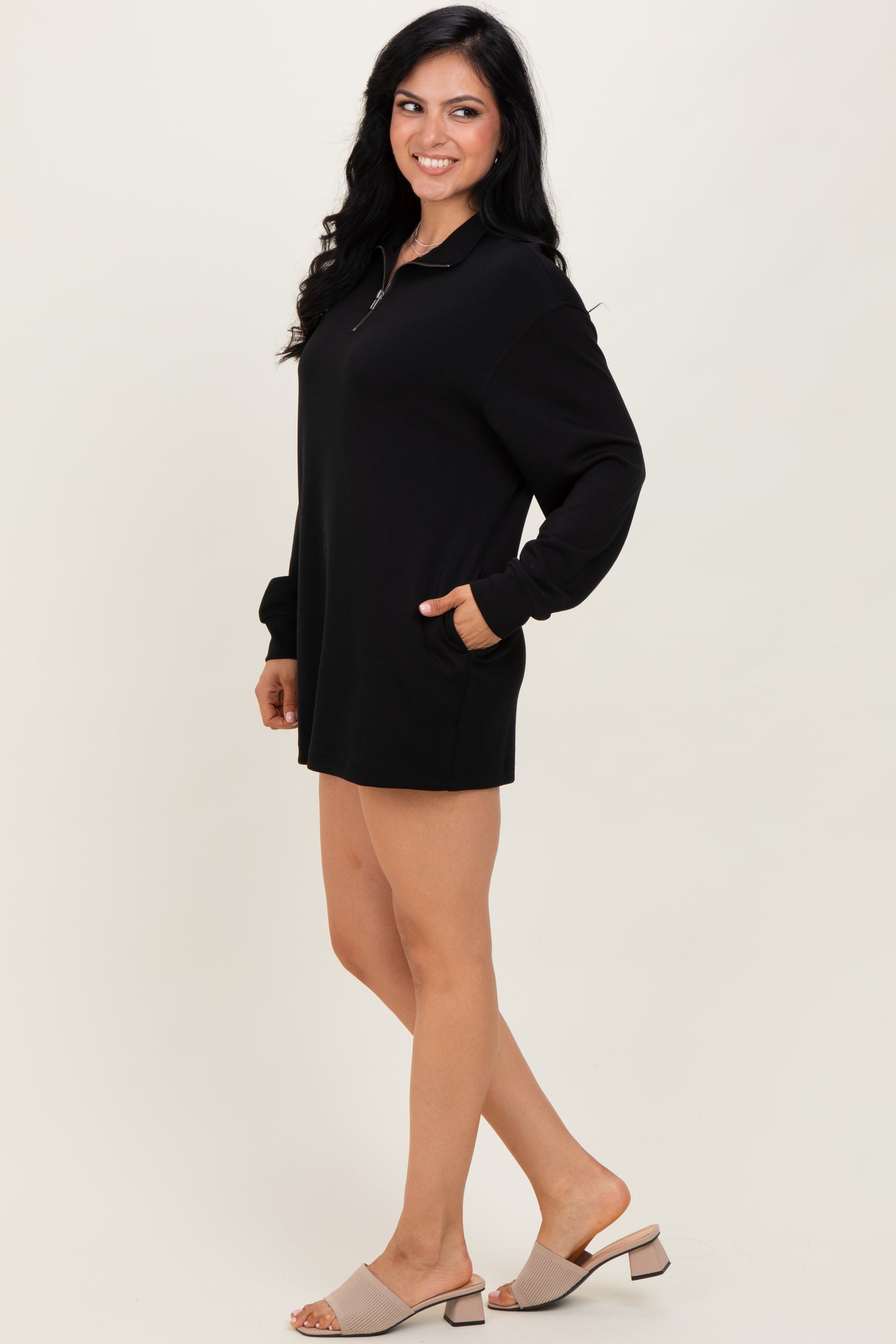 woman wearing a black oversized zip-up dress with long sleeves and pockets, standing on a neutral background