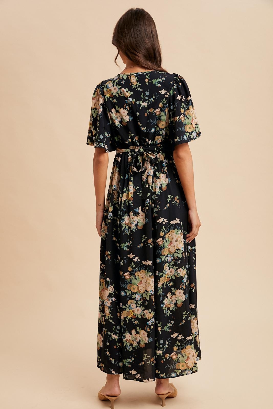 woman wearing a black floral maxi dress with short sleeves and a tied waist detail, seen from the back