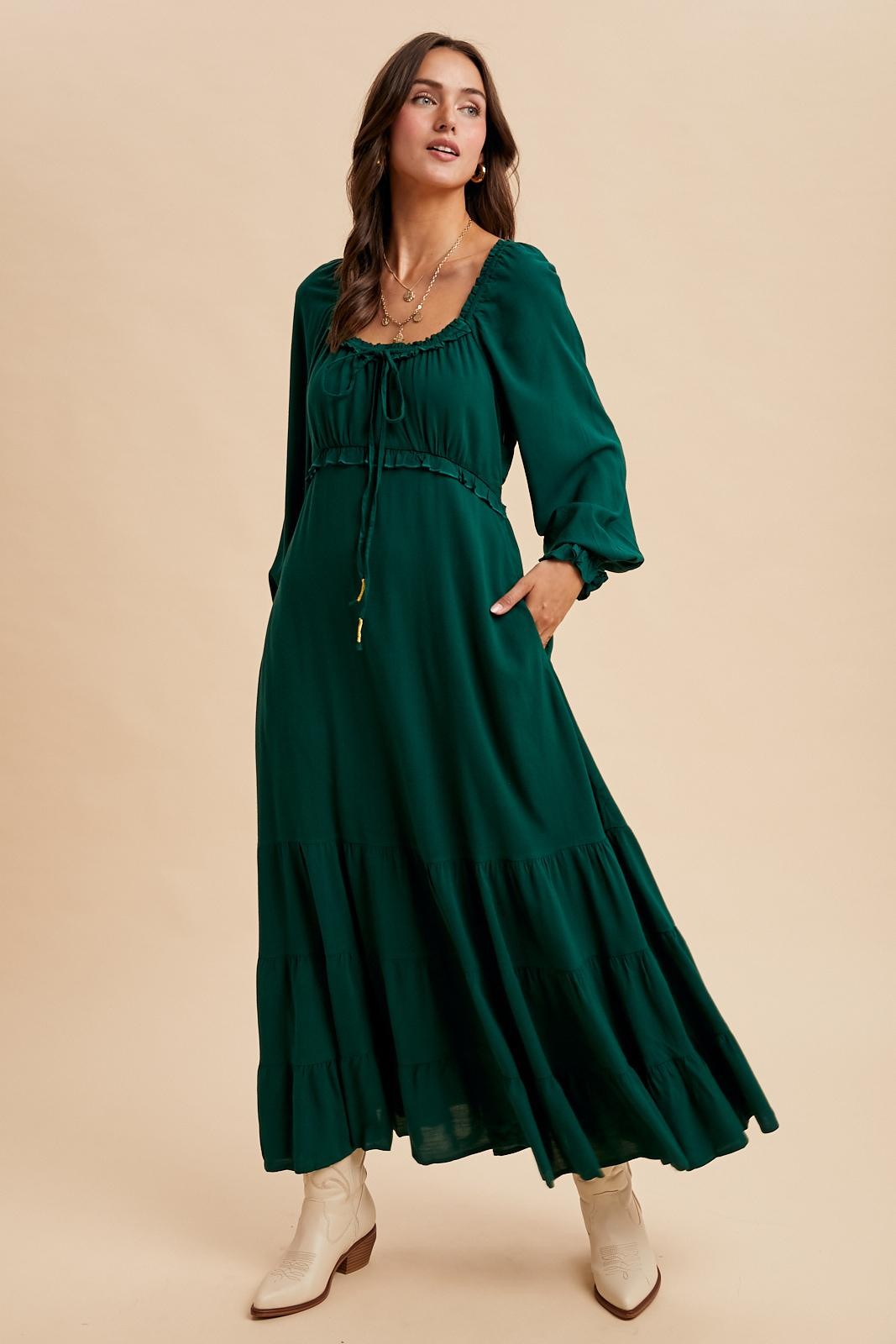 woman wearing a dark green maxi dress with puff sleeves, drawstring neckline, and tiered skirt, paired with white ankle boots