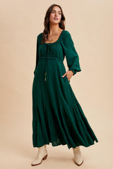 woman wearing a dark green maxi dress with puff sleeves, drawstring neckline, and tiered skirt, paired with white ankle boots