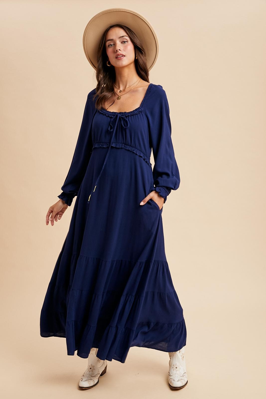 woman wearing a navy blue maxi dress with long sleeves, tie detail at the neckline, and pockets, styled with a wide-brimmed hat
