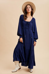 woman wearing a navy blue long sleeve maxi dress with tiered skirt and a tie neckline, paired with white cowboy boots and a wide-brim hat