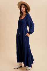 woman wearing a navy blue long sleeve maxi dress with a tiered skirt and a tie detail at the neckline, accessorized with a hat