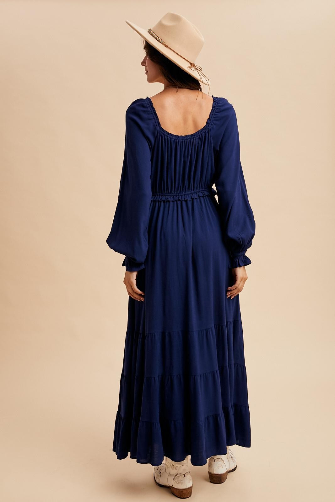 woman wearing a navy blue long sleeve tiered maxi dress with a smocked back and cinched waist, styled with a beige hat