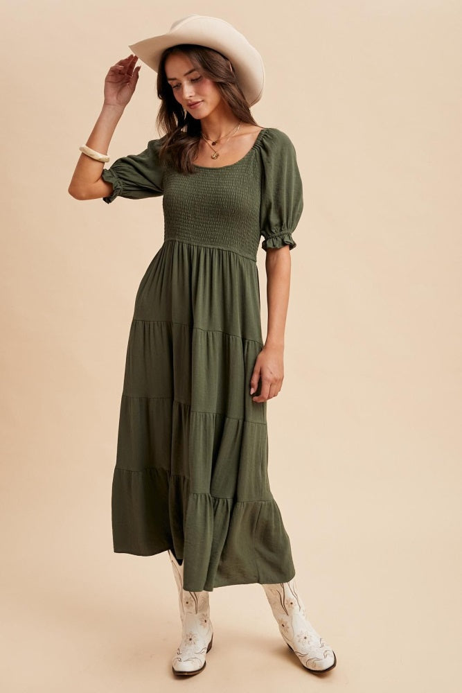 woman wearing an olive green tiered maxi dress with puff sleeves and cowboy boots, standing against a beige background