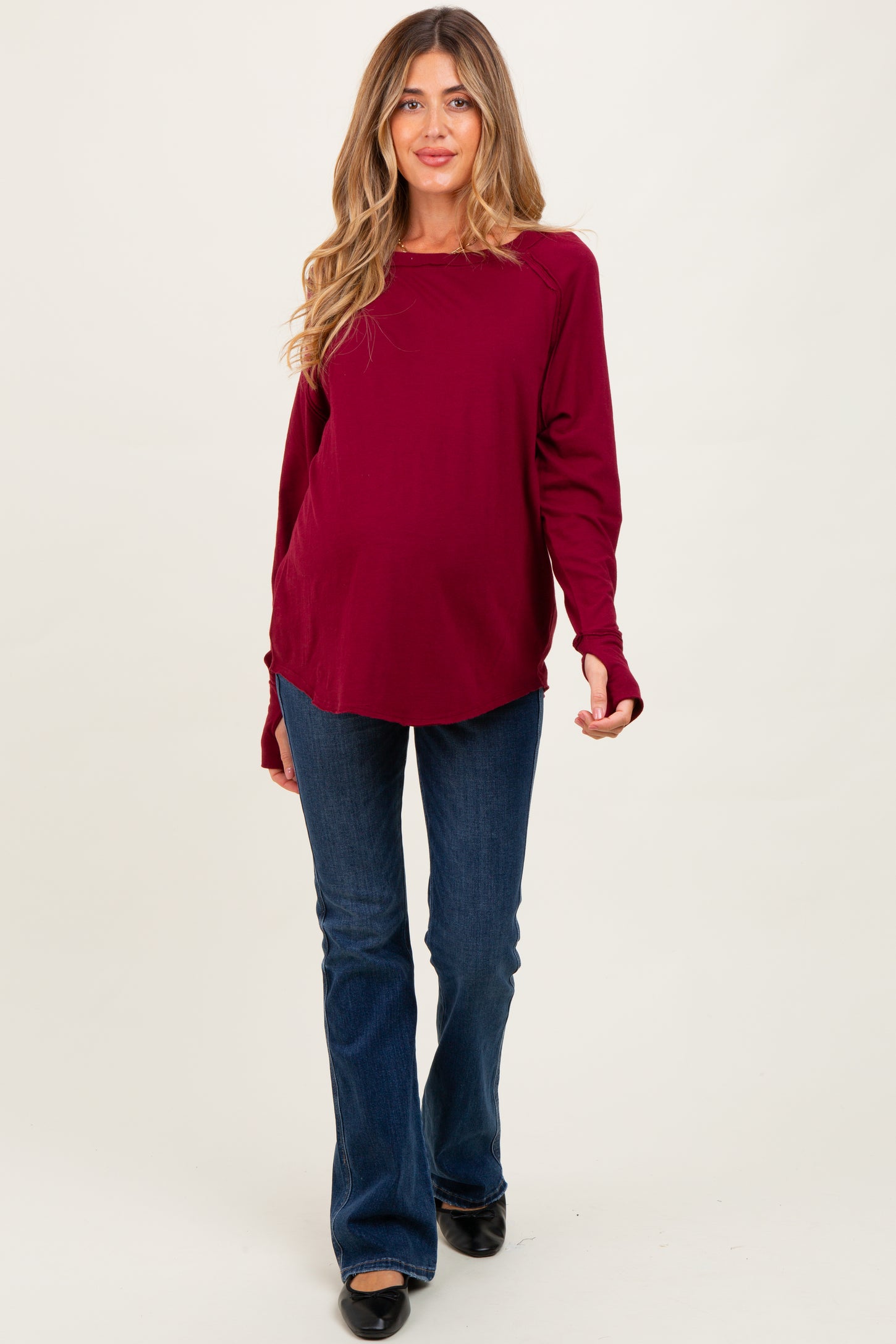 woman wearing a burgundy long sleeve maternity top and blue flared jeans, showcasing her baby bump