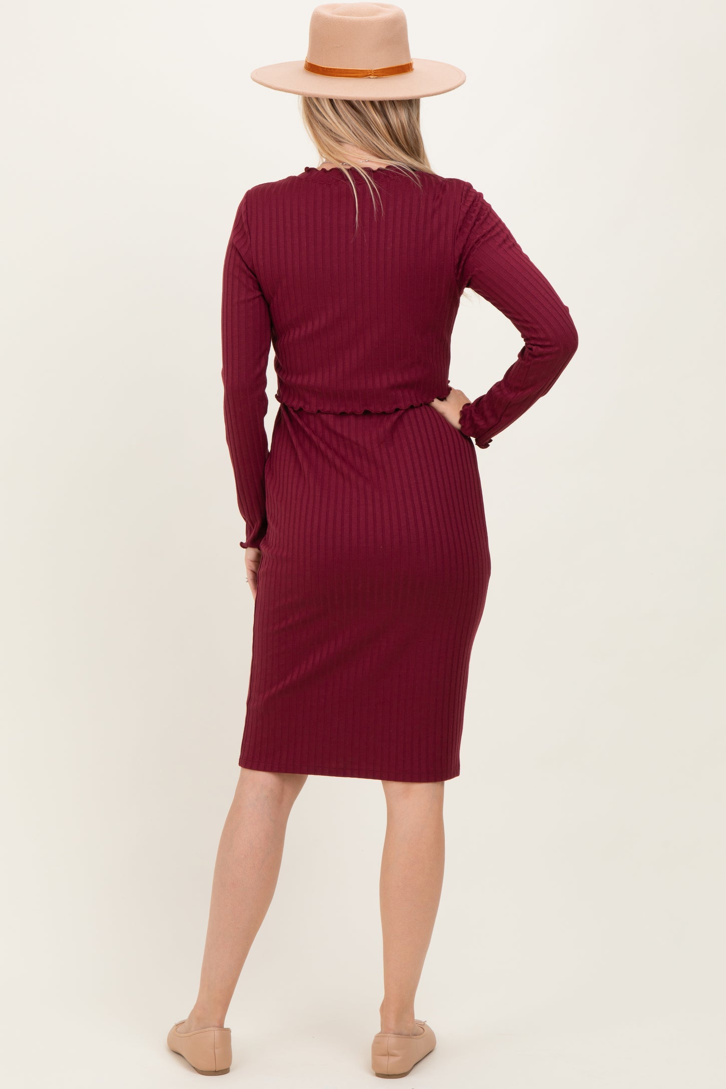 woman wearing a burgundy ribbed midi dress with long sleeves and a tie at the waist, standing with a hat on