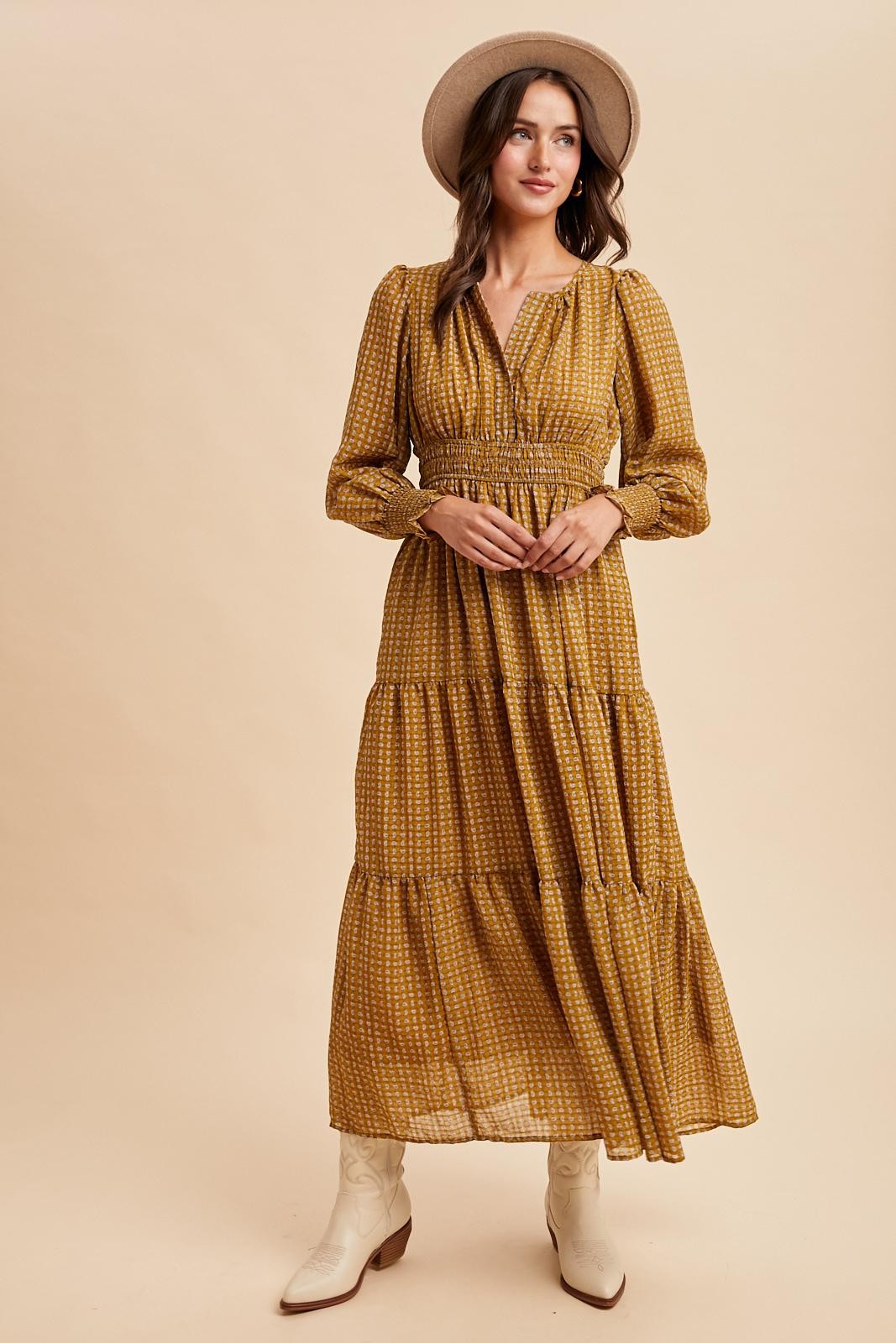 woman wearing a mustard yellow long sleeve maxi dress with a smocked waist and tiered skirt, accessorized with a tan hat and white boots