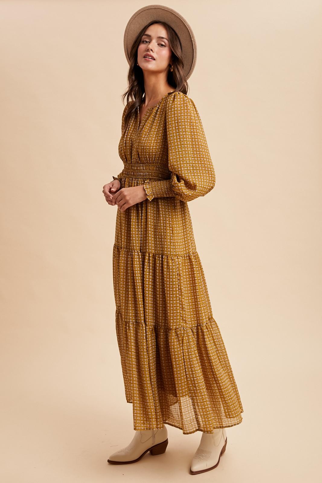 woman wearing a mustard yellow tiered maxi dress with long sleeves and a tie neckline, paired with a wide-brimmed hat and white boots