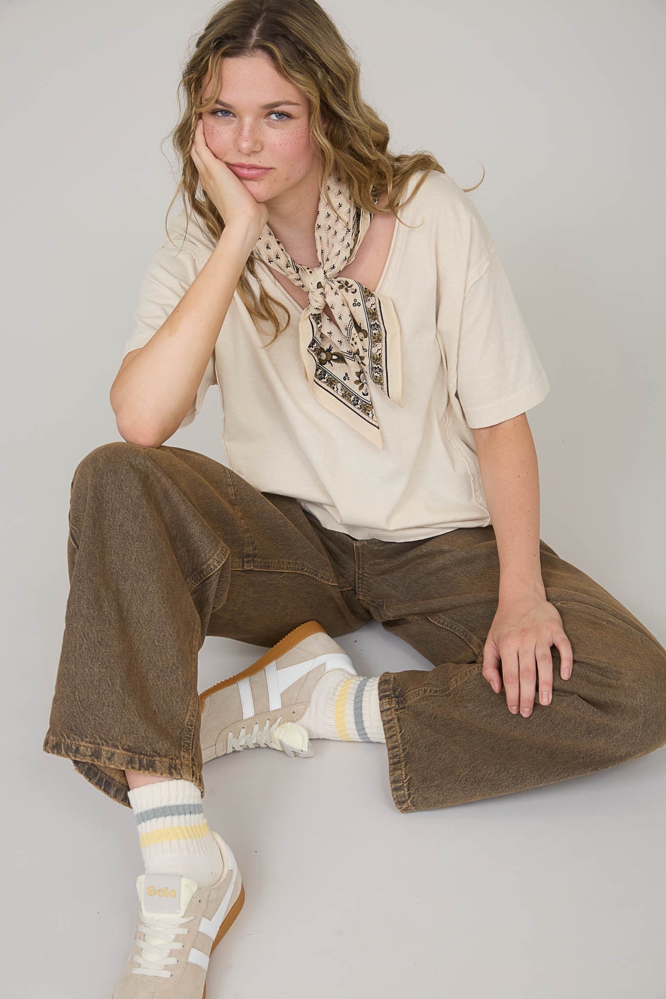 woman wearing a beige oversized t-shirt with a patterned scarf, sitting in brown wide-leg jeans and white sneakers with striped socks