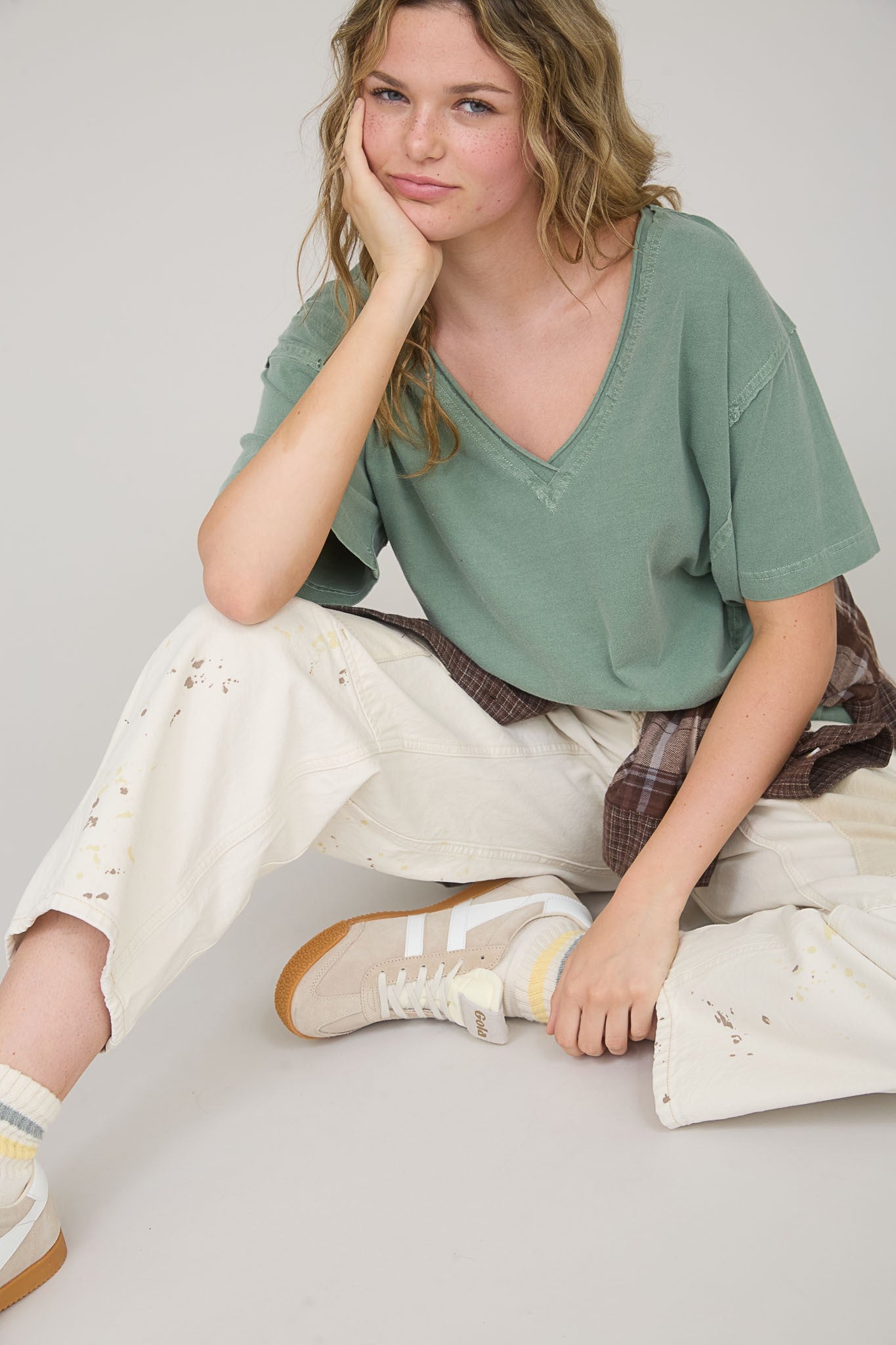woman sitting in a relaxed pose wearing a green v-neck t-shirt, off-white distressed pants, and beige sneakers
