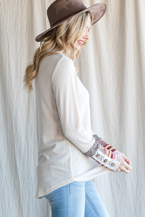 woman wearing a cream long sleeve top with patterned sleeves and a floppy hat, smiling against a neutral background