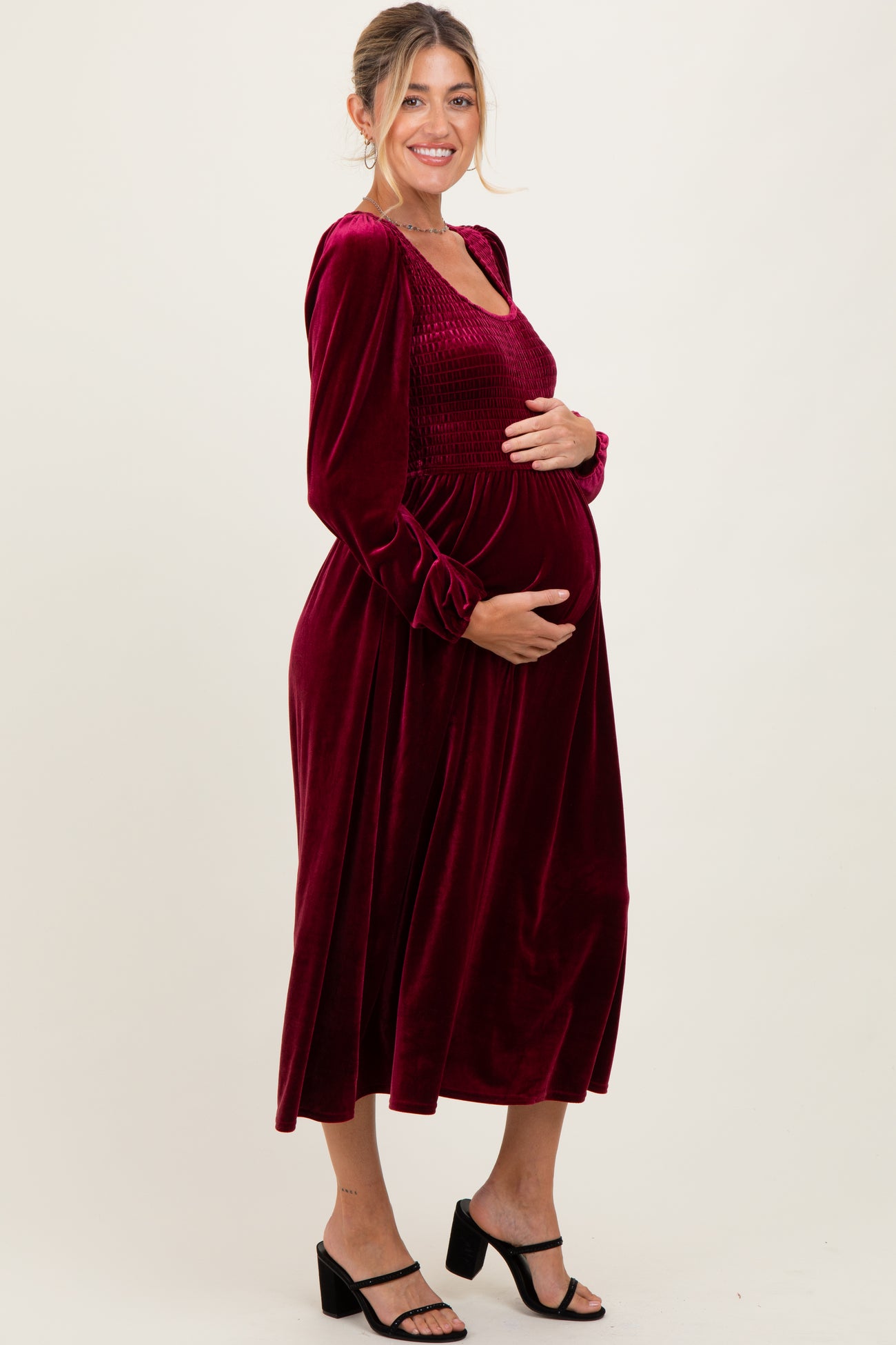 woman wearing a burgundy velvet maternity dress with long sleeves and smocked bodice, holding her baby bump