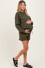 woman wearing an olive green maternity sweatshirt and matching shorts, holding her baby bump