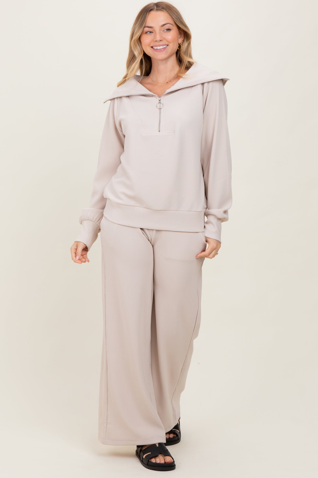 woman wearing a beige ribbed zip-up hoodie and matching wide-leg pants, smiling and standing against a neutral background