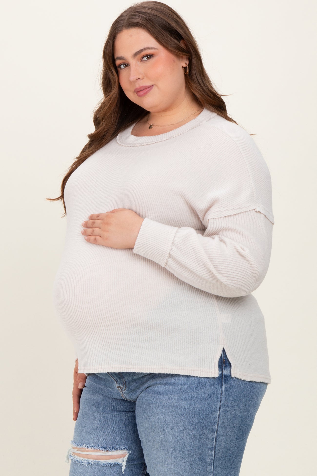 woman wearing a light beige maternity sweater with side slits and a visible baby bump, paired with blue jeans