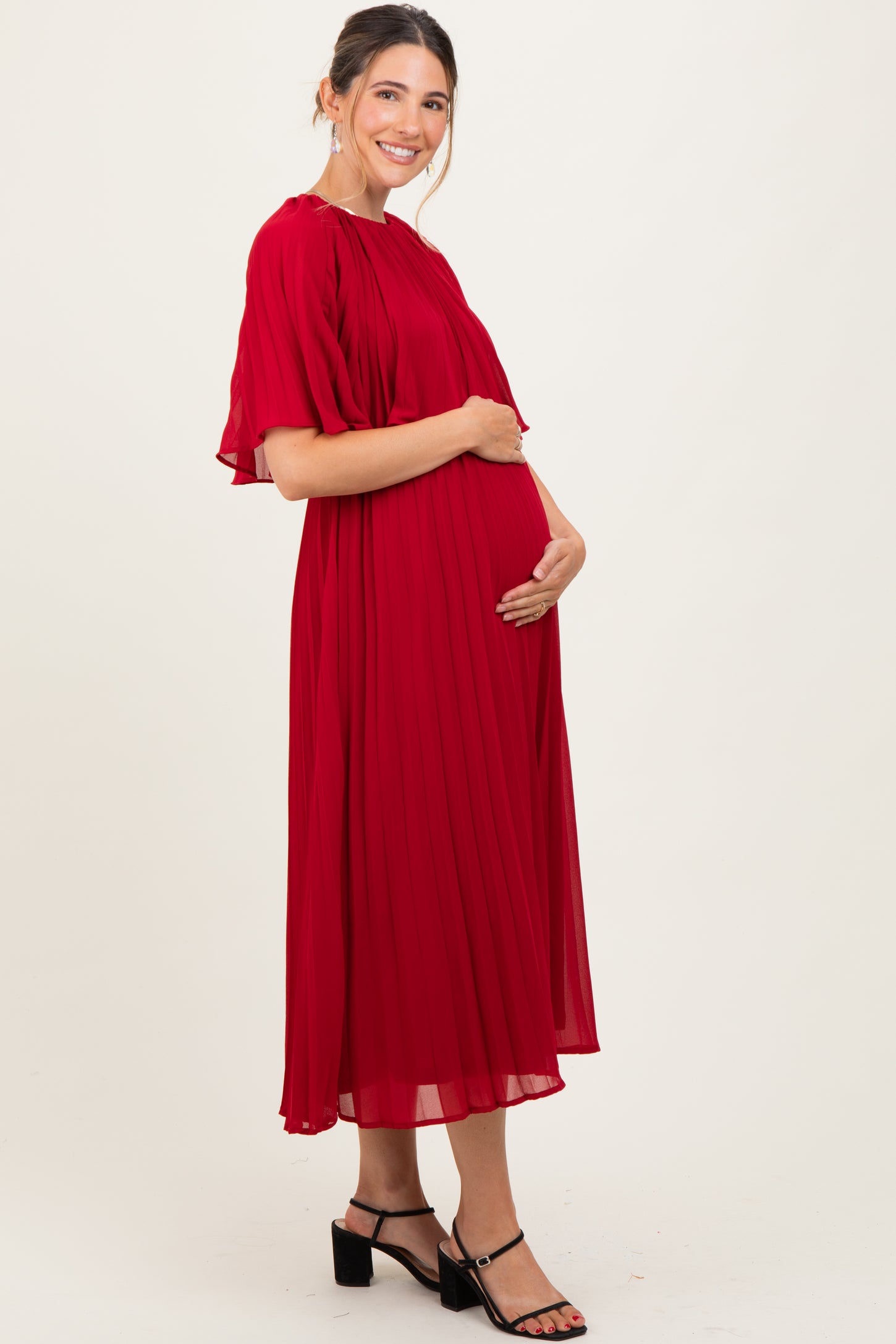 woman wearing a flowing red pleated maternity dress and cradling her baby bump