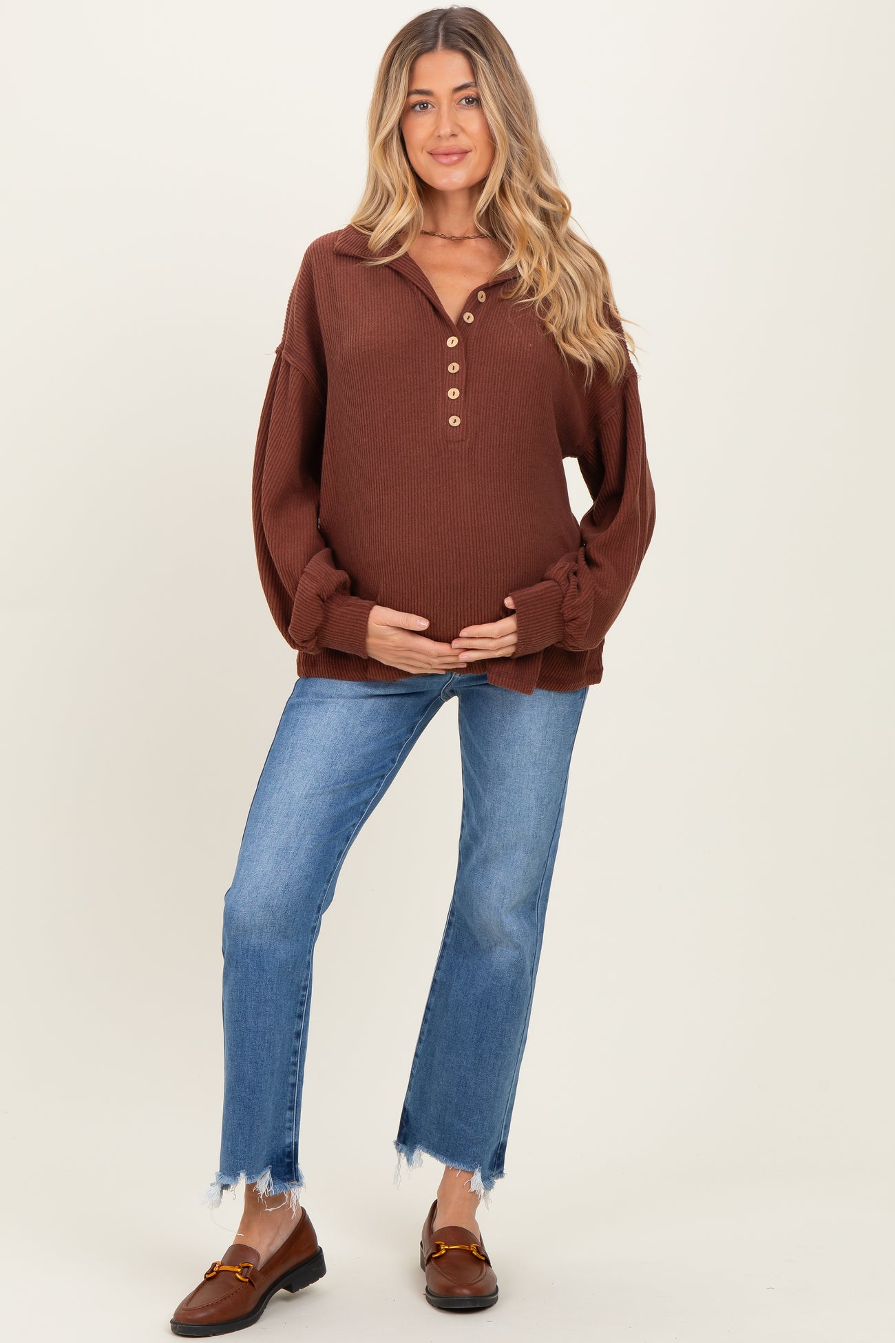 woman wearing a brown maternity pullover with button details and a baby bump, paired with light blue jeans