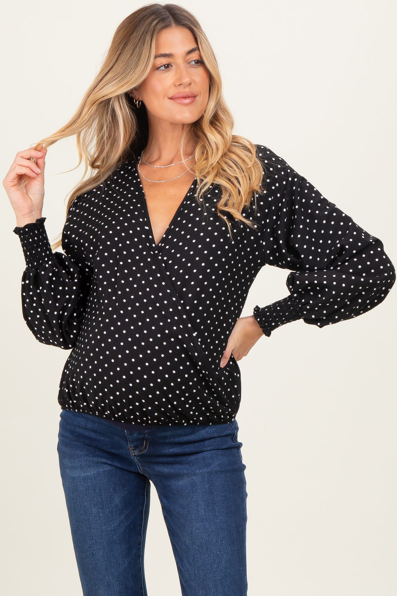 woman wearing a black maternity wrap blouse with white polka dots and puff sleeves, smiling and posing with one hand in her hair