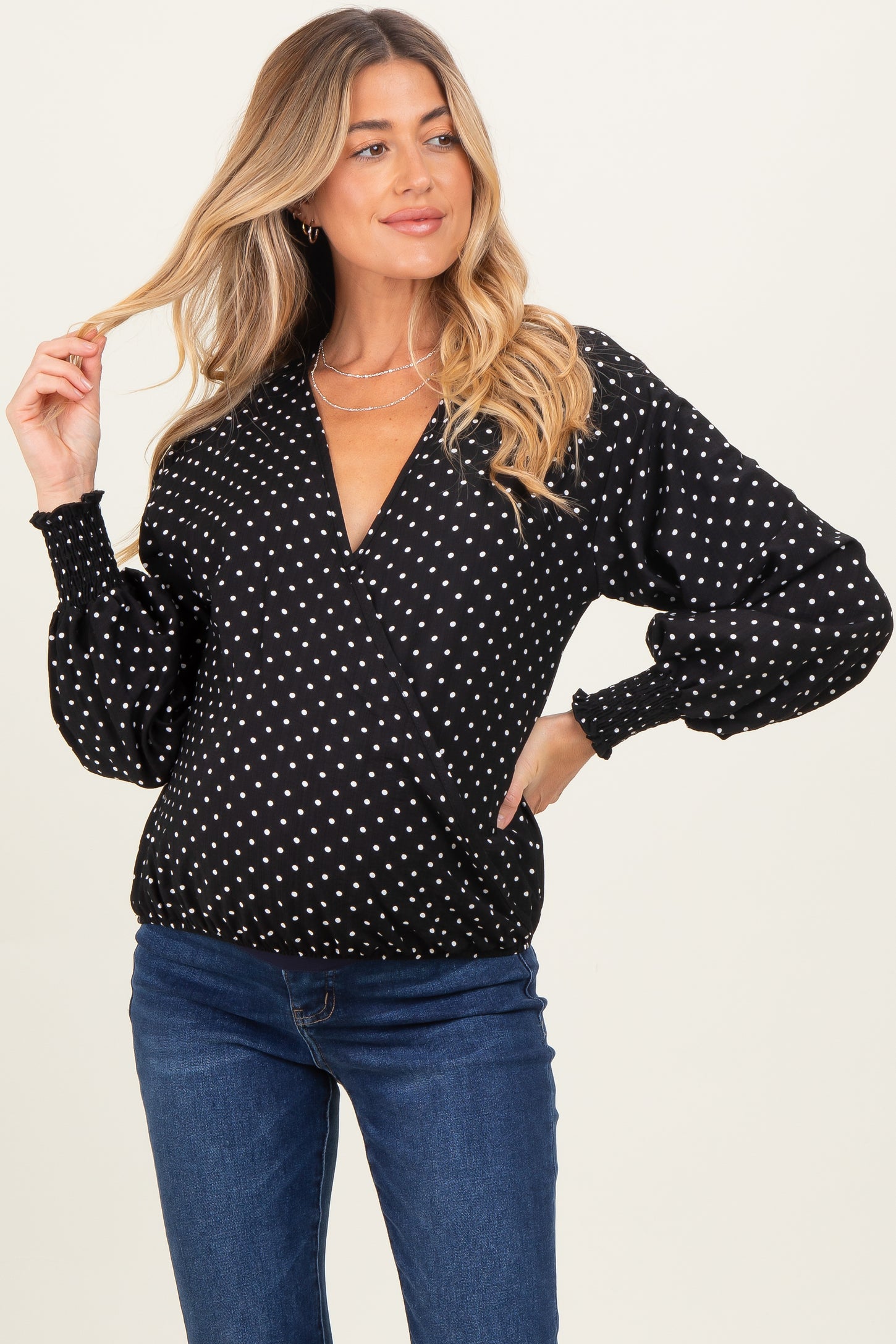 woman wearing a black maternity wrap blouse with white polka dots and puff sleeves, smiling and posing with one hand in her hair