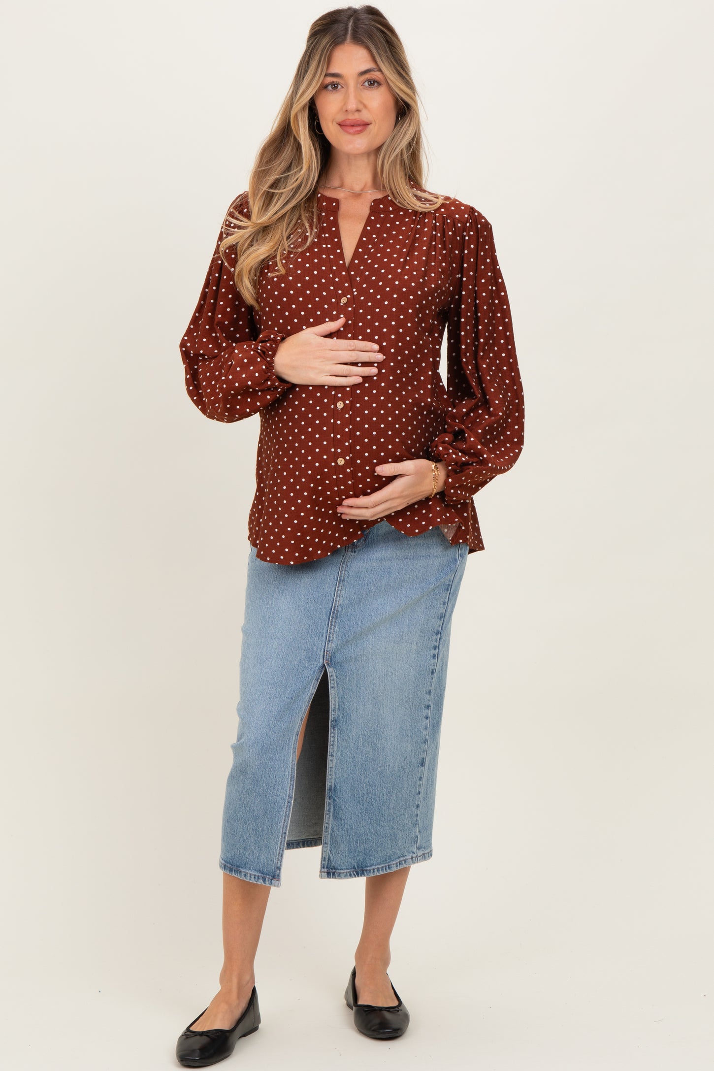 woman wearing a burgundy polka dot maternity top and a denim skirt, gently holding her baby bump