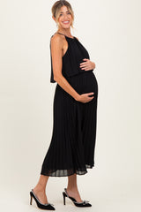 woman wearing a black pleated maternity dress with a flowy overlay and holding her baby bump