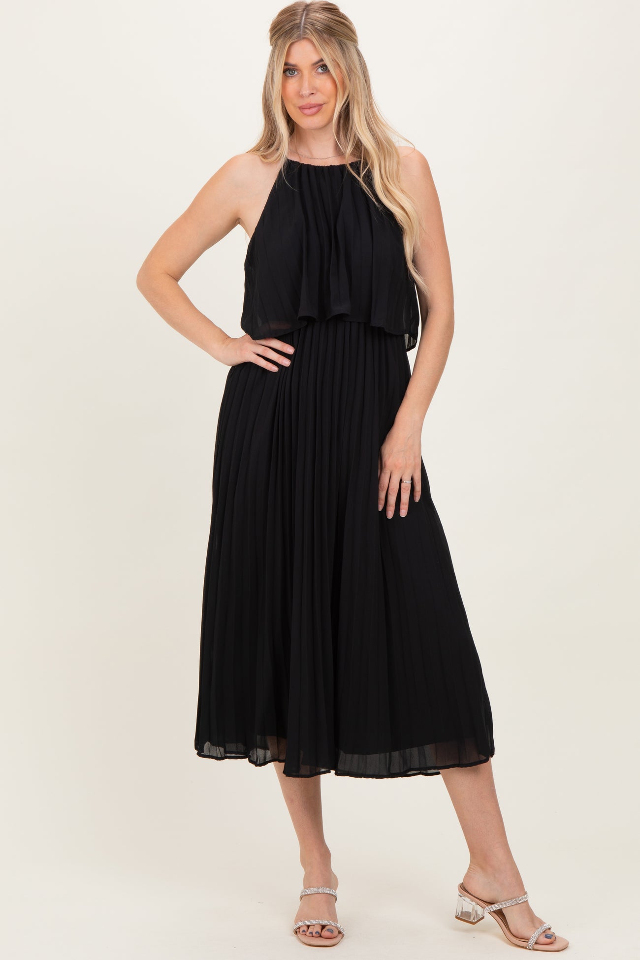 woman wearing a black pleated maxi dress with a halter neck and layered top, standing with one hand on her hip