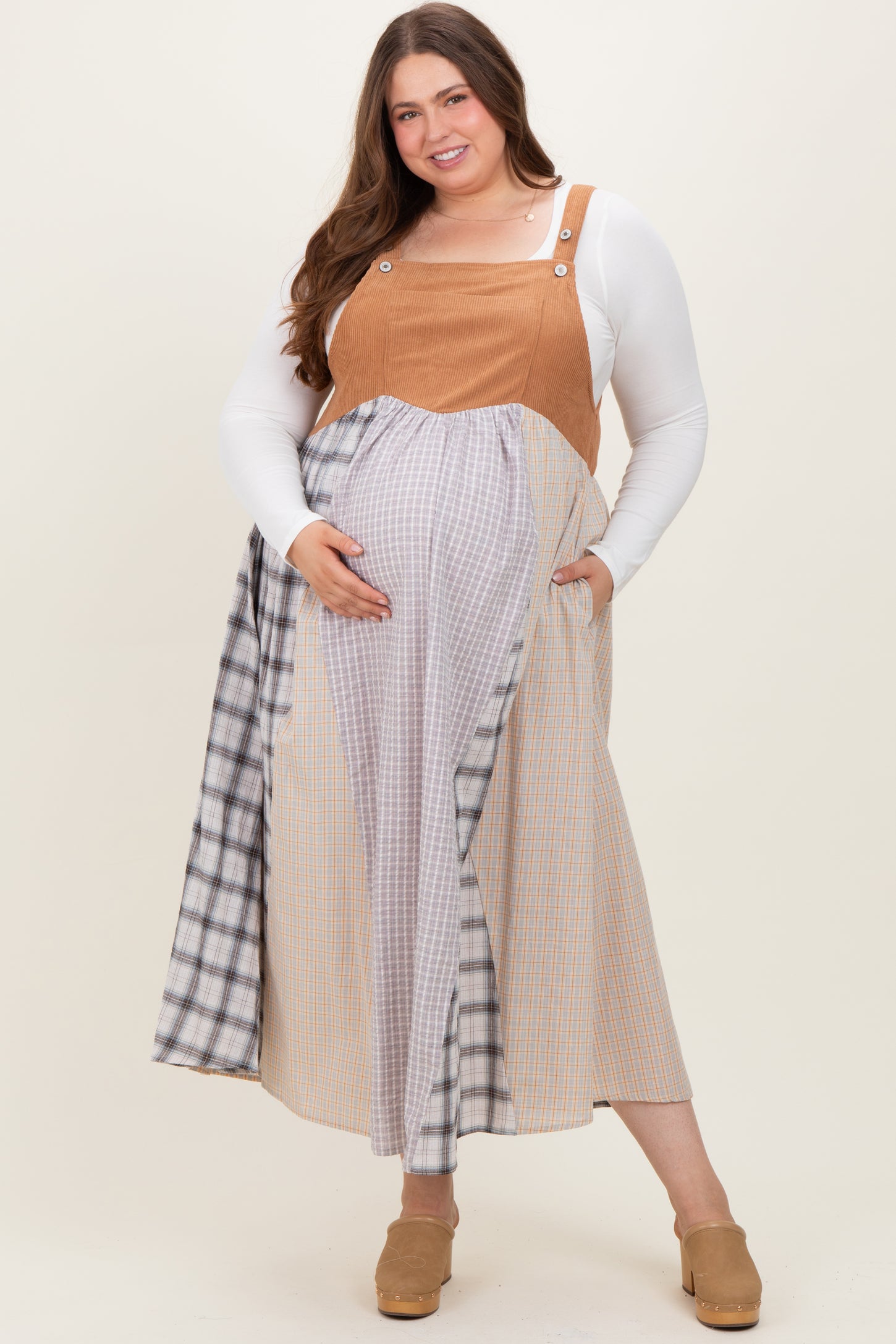 woman wearing a tan maternity plus overall dress with plaid detailing, showing her baby bump and smiling