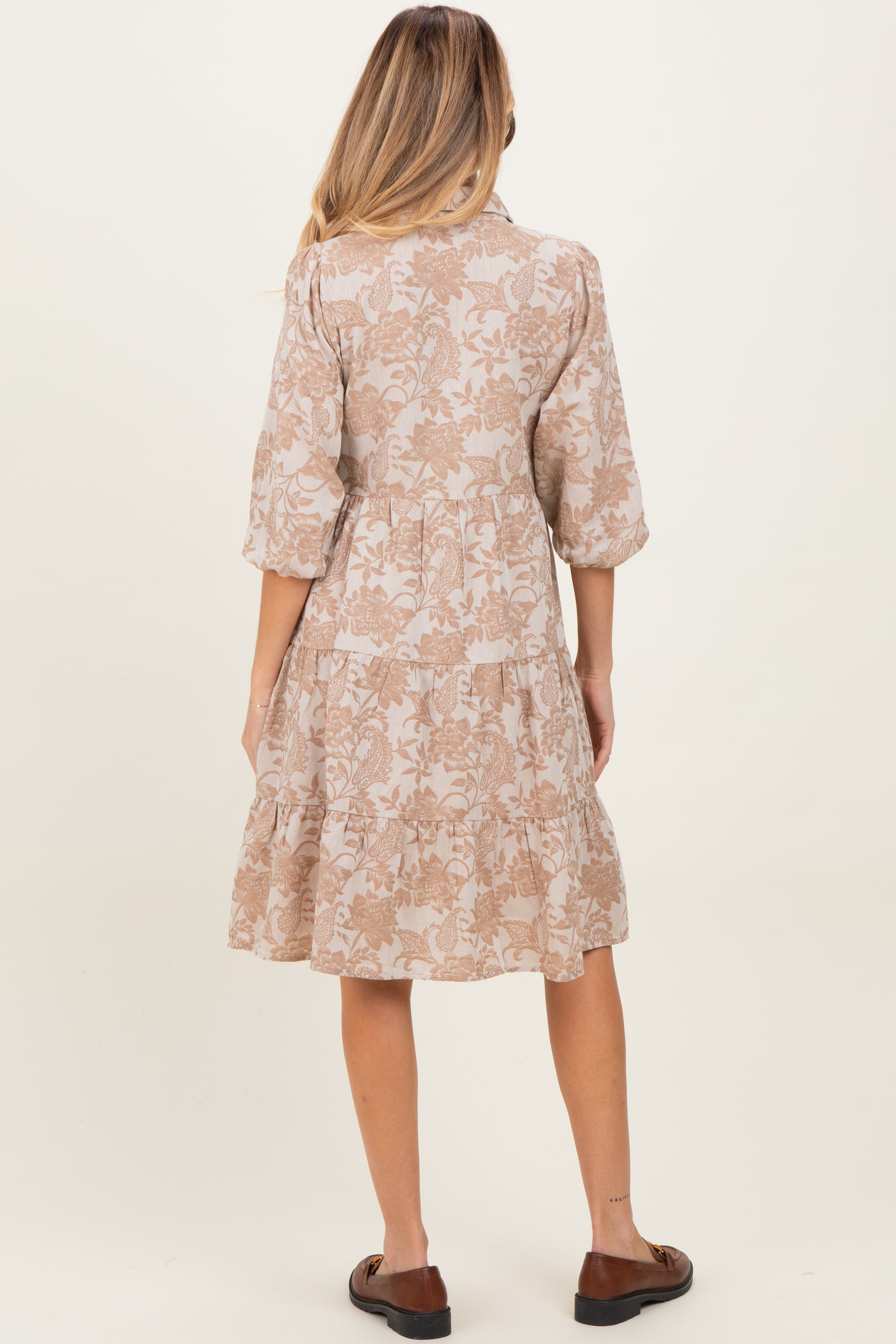woman wearing a beige floral maternity dress with three-quarter sleeves and a tiered skirt, standing with her back to the camera
