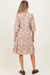 woman wearing a beige floral maternity dress with three-quarter sleeves and a tiered skirt, standing with her back to the camera