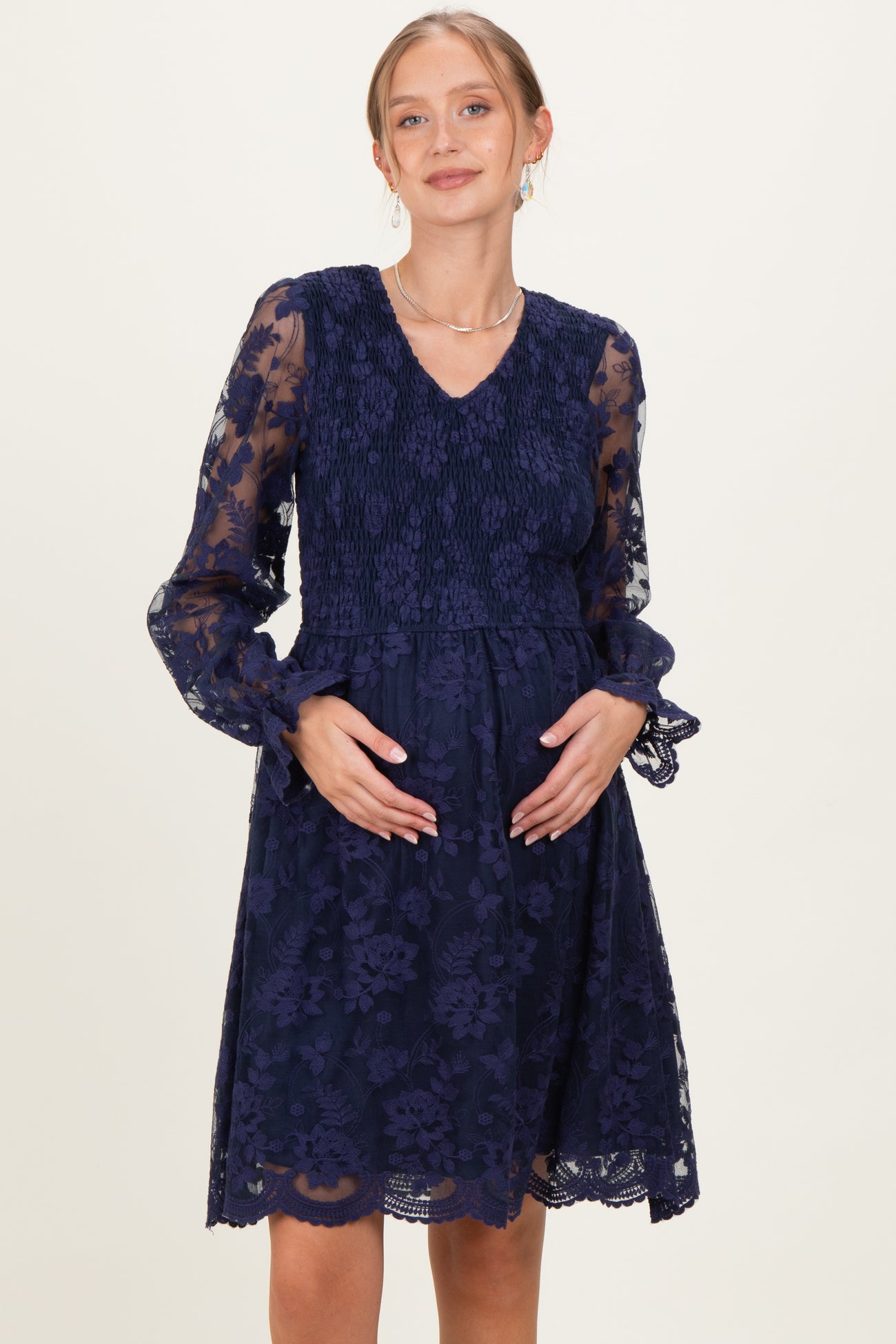 woman wearing a navy blue lace maternity dress with smocked bodice and long sleeves, holding her baby bump