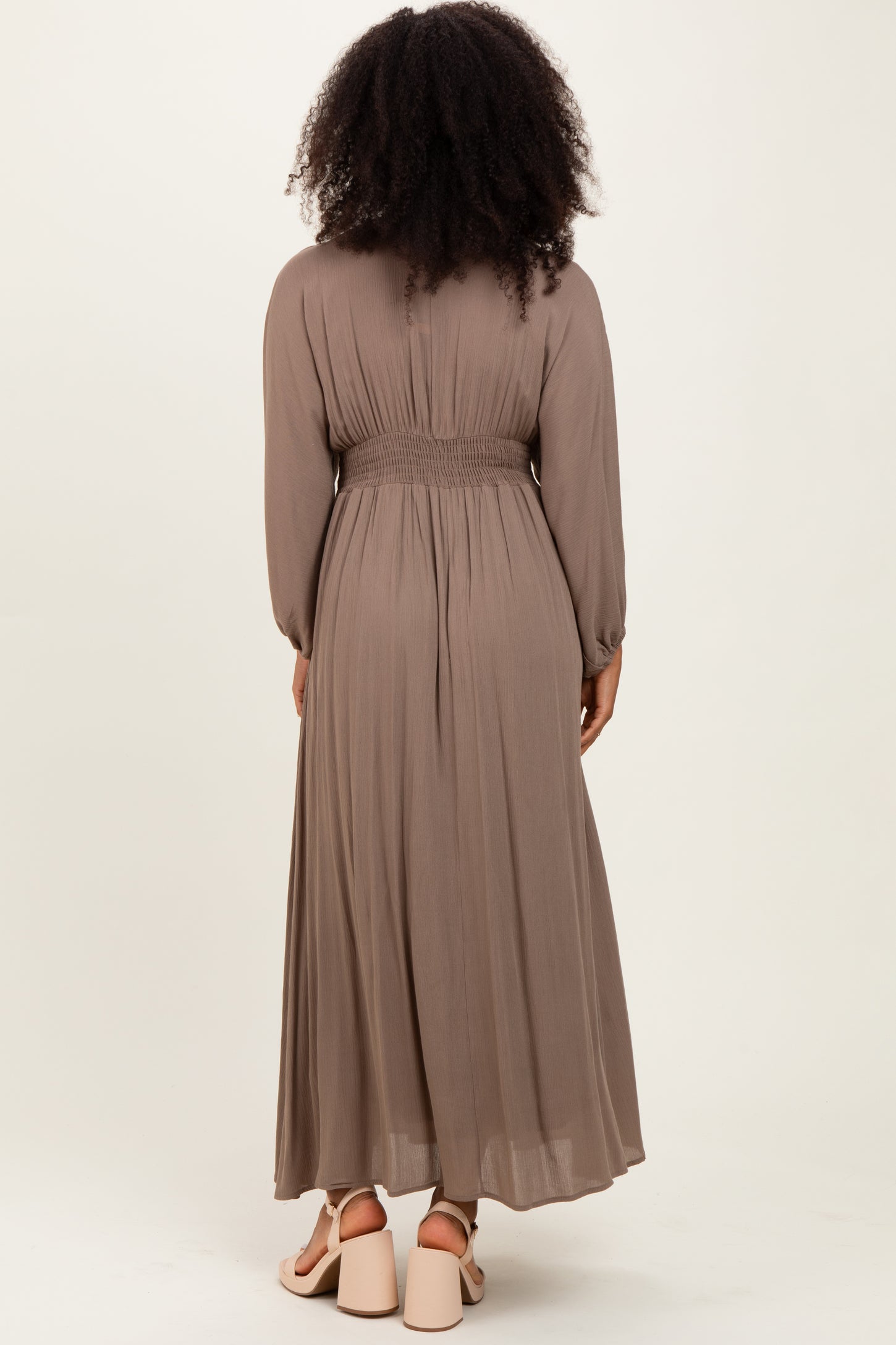 woman wearing a long taupe maxi dress with a smocked waist and long sleeves, viewed from the back