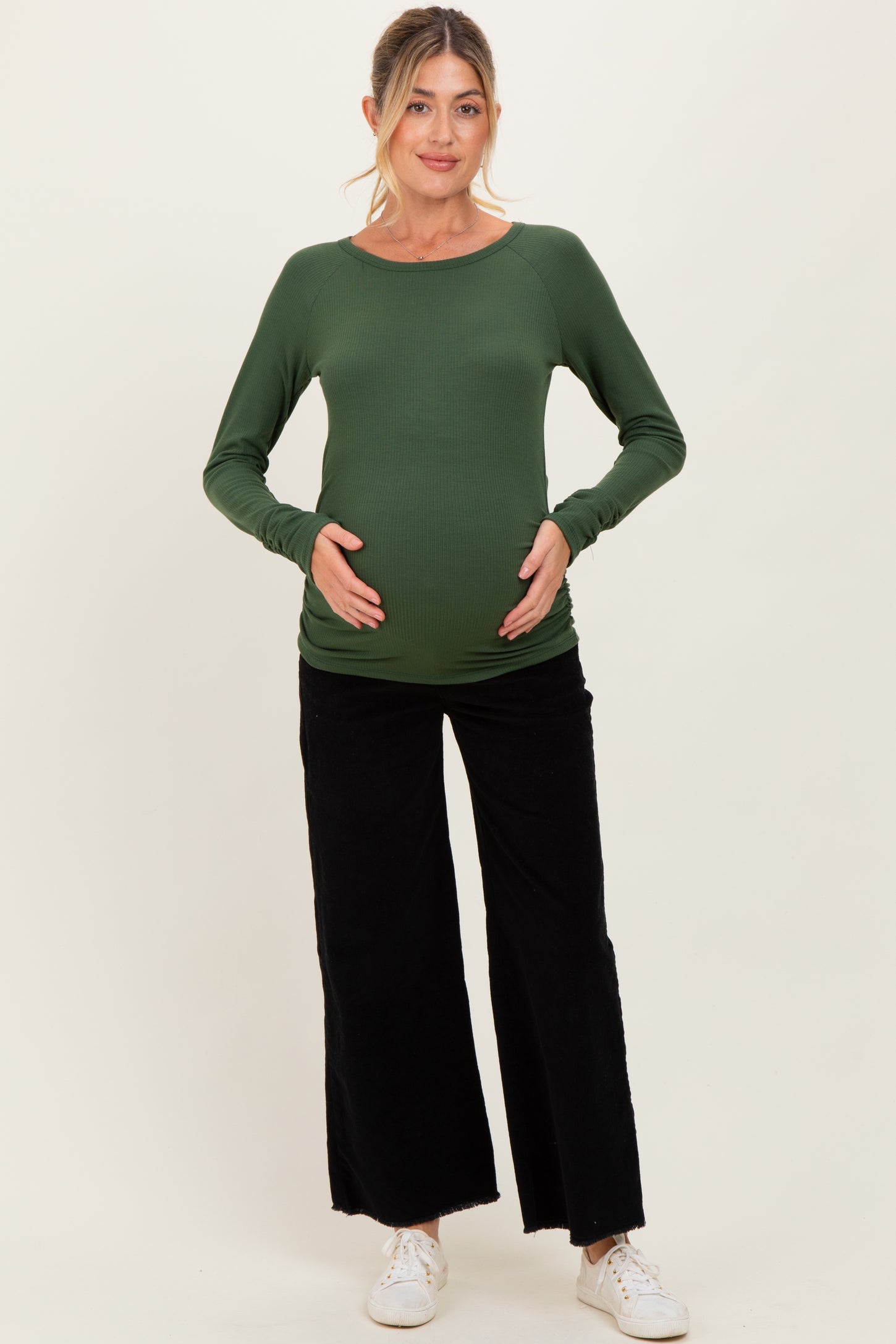 woman wearing a dark green maternity long sleeve top and black wide-leg pants, holding her baby bump